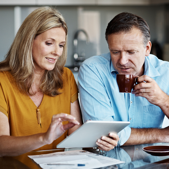 Middle aged man and woman drinking coffee at home while looking at a tablet device