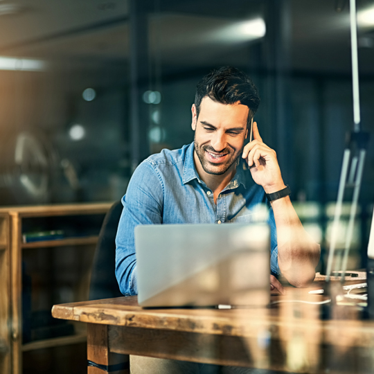 Man smiling and talking on a cell phone while looking at a laptop