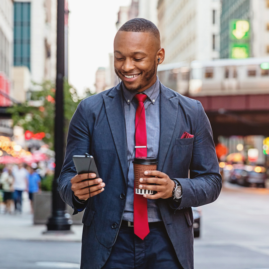 Man in business attire walking down the street smiling, holding coffee, looking at a smartphone