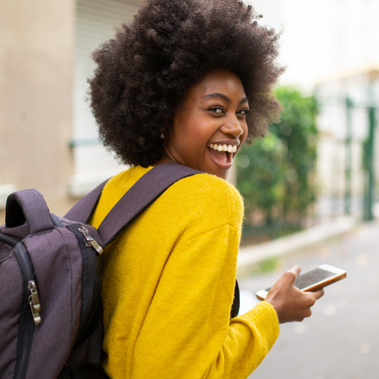 College student looking over her shoulder smiling with a backpack and smartphone