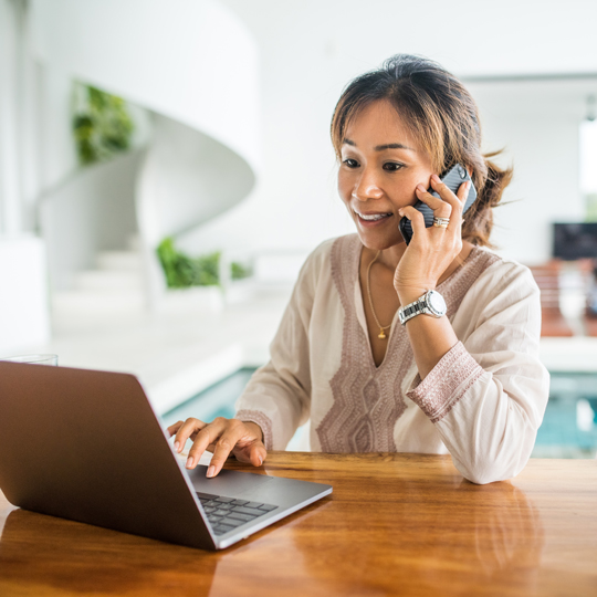 Woman smiling and working on a laptop while talking on a smartphone